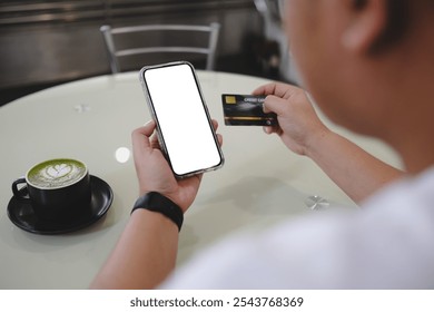 Person making digital payment in cafe setting, holding smartphone with blank screen and credit card, with matcha latte art coffee in black cup on white table. - Powered by Shutterstock