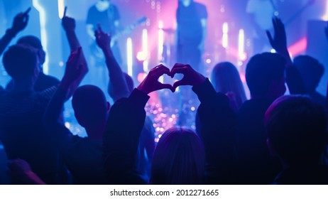 Person is Making a Heart Sign Gesture and Holding Hands Up at a Performance. Rock Band Playing a Song at a Concert in a Night Club on Stage with Bright Colorful Strobing Lights. - Powered by Shutterstock