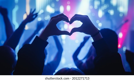 Person is Making a Heart Sign Gesture and Holding Hands Up at a Performance. Rock Band Playing a Song at a Concert in a Night Club on Stage with Bright Colorful Strobing Lights. - Powered by Shutterstock