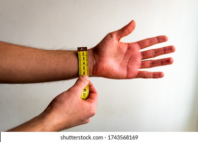 Person Measuring The Wrist Of His Left Arm / Hand With A Yellow Tape Measure. White Background. Olive Skin Tone. 