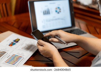 Person multitasking, scrolling smartphone while reviewing business data on a laptop and reports
