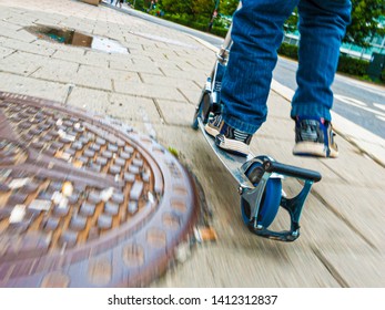 Person On A Kickbike Passing A Rusty Manhole Cover.