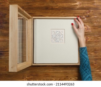 person opens a photobook in wooden box with glass lid. Family photoalbum on the table with copy space for text. womans hand holding and looking  stylish wedding photo album with decor shield 