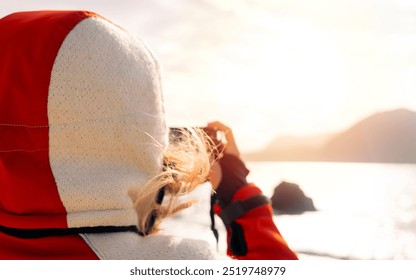 Person in an orange and white hooded jacket, holding a camera and taking a photo, possibly of a scenic view or sunset over water and mountains. The focus is on the person’s back, with the sunlight  - Powered by Shutterstock