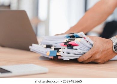 Person organizing stack of documents with colorful paper clips on wooden desk near laptop.