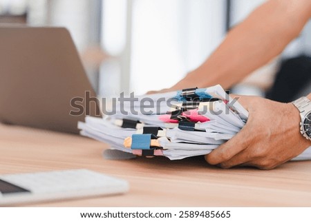 Person organizing stack of documents with colorful paper clips on wooden desk near laptop.