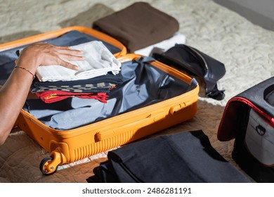 A person packing clothes into an open orange suitcase on a bed. Various items of clothing and a black toiletry bag are visible. - Powered by Shutterstock