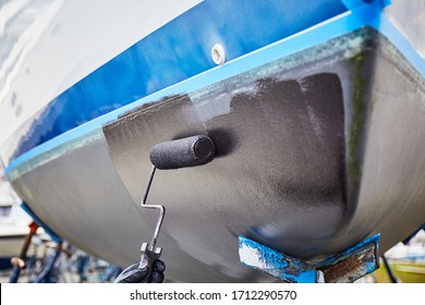 Person painting a boat's bottom with a fresh paint, using roller and black paint, outdoors in the boatyard