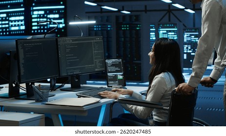 Person with paraplegia helped by coworker in data center to arrive at desk, coding on PC. Woman in wheelchair doing maintenance on server room infrastructure rackmounts, camera A