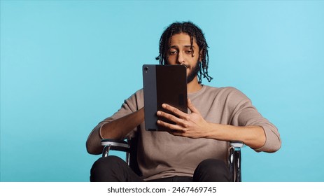 Person with paraplegia using tablet to post images on social media, doing selfie and writing photo caption. Man in wheelchair using online editing application on digital device, studio, camera A