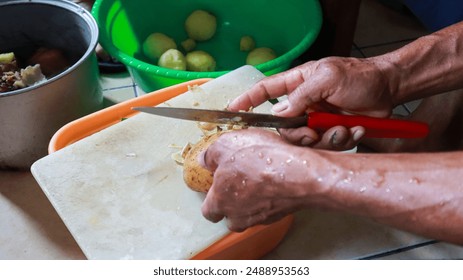 A person is peeling a potato with a knife on a cutting board. A Man peels potatoes with a sharp knife. - Powered by Shutterstock