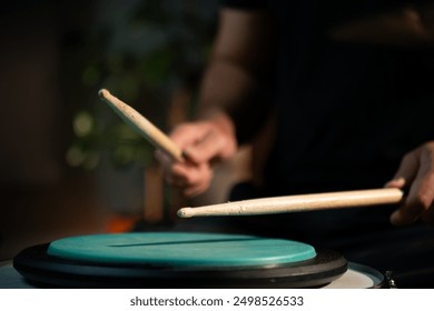 A person plays drumsticks on a green practice pad while sitting in a dimly lit studio. The pad is on top of a drum and the person is wearing a black shirt - Powered by Shutterstock