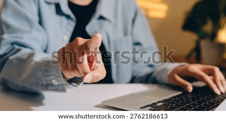 Person pointing finger at camera while using laptop on desk, showing focus and engagement in digital work or communication