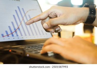 A Person pointing at a line graph on a laptop screen, with a bar chart visible in the background. The person is wearing a black wristwatch and is typing on the keyboard. - Powered by Shutterstock