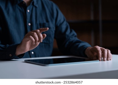 A person points to a tablet on a white desk. The low-light setting creates a moody atmosphere, suggesting concentration or focus - Powered by Shutterstock