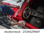 A person polishes the shiny surface of a vintage red sports car in a garage. Sunlight highlights the car