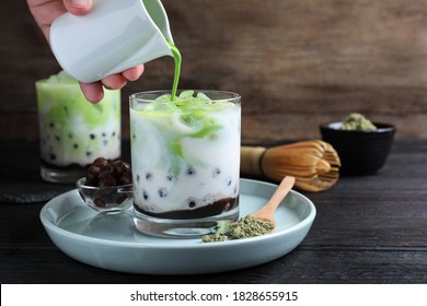 Person pouring green matcha into glass with milk bubble tea at black wooden table, closeup