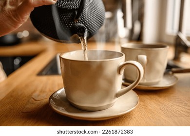 Person pouring hot water from kettle in cup at kitchen to prepare tea. Teapot and ceramic mug with warm beverage - Powered by Shutterstock