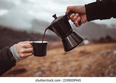 A Person Pouring Out Coffee From Moka Pot Into A Cup Outdoors In Front Of Foggy Mountains Background