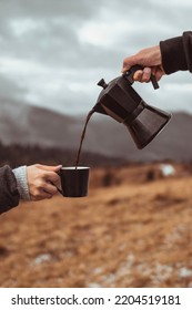 A Person Pouring Out Coffee From Moka Pot Into A Cup Outdoors In Front Of Foggy Mountains Background