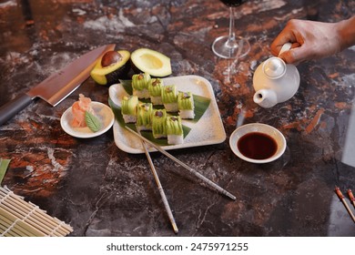 person pouring soy sauce from a small ceramic jug into a small white bowl, with a plate of sushi rolls in the background - Powered by Shutterstock