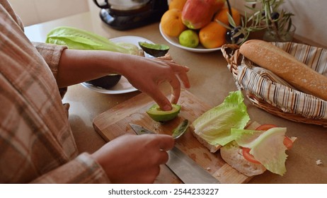 A person prepares a fresh sandwich with sliced avocado, lettuce, and tomato on a wooden board. Nearby are fruits and baguette, conveying a healthy lifestyle. - Powered by Shutterstock