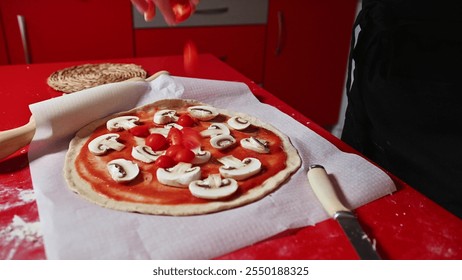 A person prepares a homemade pizza with mushrooms and cherry tomatoes on a red countertop. The kitchen setup and fresh ingredients highlight a cozy cooking atmosphere. - Powered by Shutterstock