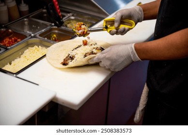 A person is preparing a burrito by adding ingredients to a tortilla while wearing gloves in a kitchen setting. - Powered by Shutterstock