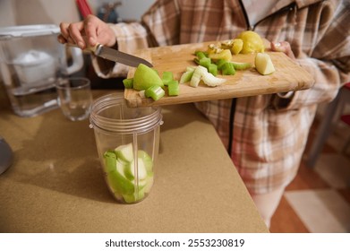 A person is preparing a nutritious green juice by cutting fresh apples and celery. The ingredients are placed in a blender cup on the kitchen counter. Emphasis on healthy eating. - Powered by Shutterstock