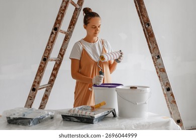 A person is preparing to paint a wall using a roller. They stand near a ladder, surrounded by buckets and paint supplies in a bright, airy space.