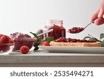 Person preparing toast with raspberry jam on white plate for breakfast at kitchen table with jars full of jam and bowl with berries. Front view.