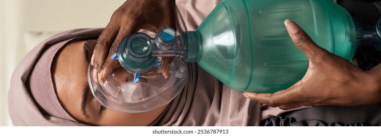 Person providing CPR by using breathing apparatus, focusing on emergency aid. Hands holding mask firmly against Muslim patient's face for effective ventilation - Powered by Shutterstock