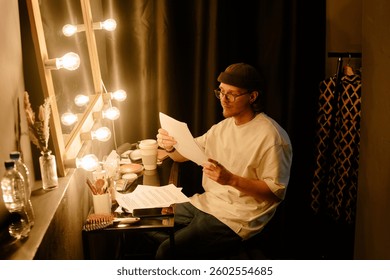 Person reading script while seated beside an illuminated vanity mirror inside dressing room, creating a focused ambiance with a touch of anticipation
