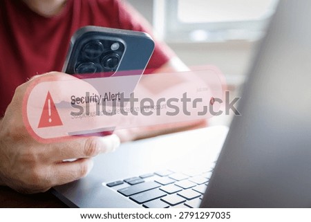 Person receiving security alert notification on smartphone about suspicious data activity, account breach, or phishing attempt while using laptop