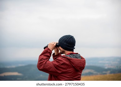 Person in a red jacket and beanie using binoculars, overlooking a vast landscape under a cloudy sky.