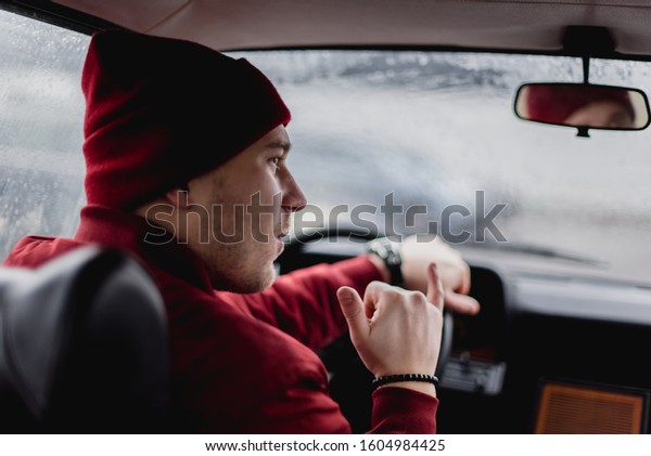 Person in red jacket sitting in vintage car with\
serious face. Concept of traveller. Person travel by car. Young guy\
before race on track.