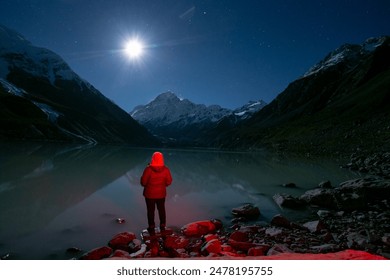 A person in a red jacket stands at the edge of a tranquil mountain lake under a starry night sky, with snow-capped peaks reflected in the calm water. The full moon casts a serene glow over landscape - Powered by Shutterstock