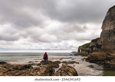 A person in a red jacket stands on rocky terrain, gazing at the ocean under a cloudy sky, with dramatic cliffs and a sea arch in the background. - Powered by Shutterstock