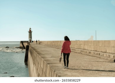 A person in a red jacket is strolling down a stone pier towards a lighthouse under a clear sky. The Douro River meets the ocean, creating a picturesque setting in Porto