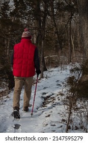 Person With A Red Puffy Vest And Walking Stick Hiking Along A Park Trail Through A Deciduous Forest During Winter In The Midwest With Snow Cover, Foot Prints, And Bare Trees And Bushes.