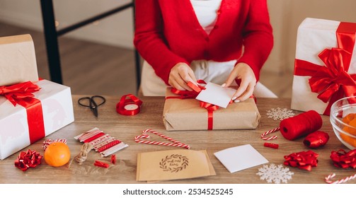 A person in red wrapping Christmas gifts on a wooden table surrounded by festive decorations, candy canes, and ribbons. The scene evokes warmth and holiday spirit. - Powered by Shutterstock