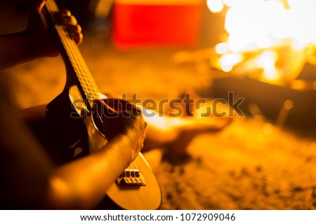 A person relaxing while sitting next to a campfire on a beach, playing a guitar