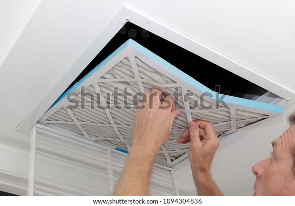 Person removing an old\
dirty air filter from a ceiling intake vent of a home HVAC system.\
Unclean gray square furnace air filter being taken out of a ceiling\
air vent.