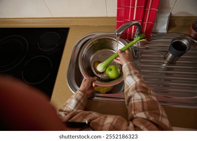 A person rinses fresh vegetables and fruits, including an apple and celery, under running water in a modern kitchen sink, emphasizing healthy cleaning habits and food hygiene. - Powered by Shutterstock