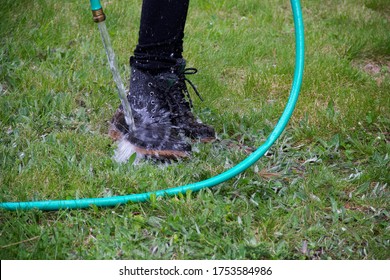 Person Rinsing Off Muddy Work Boots In Grass With Hose, Water