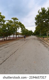 Person Running Alone On The Asphalt In The Empty Park Surrounded By Trees, On A Cloudy Day Early In The Morning.