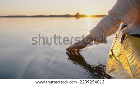 Person sails on kayak touching water with hand making small waves closeup. Anonymous enjoys canoeing and tourism. Sun rises on horizon