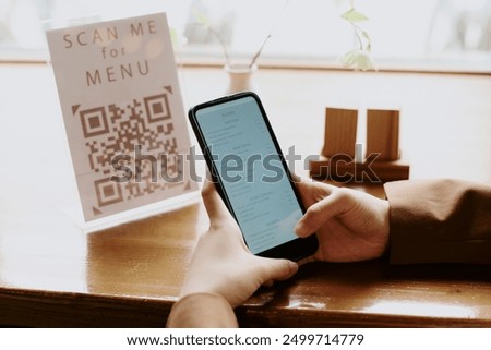 Person scanning menu QR code with smartphone in cozy restaurant setting, holding phone steadily while focusing on screen, ambient light reflecting on wooden table surface