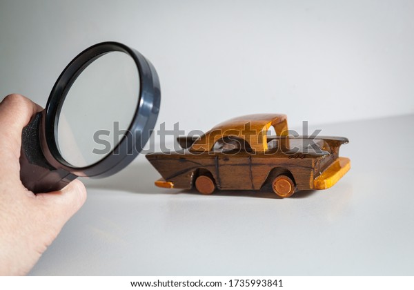 Person Scrutinizing A Wooden\
Car Model Using Magnifying Glass. Car search. Vehicle inspection\
with a magnifying glass. A man looks at a toy car with a magnifying\
glass.