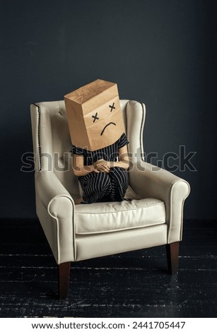 A person is seated in a chair with a paper bag covering their head, obscuring their face.